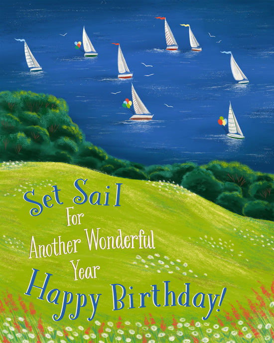 A colorful illustrated birthday card featuring a coastal seascape with sailboats on deep blue water, green hills, and white flowers. Large decorative text reads, 'Set Sail For Another Wonderful Year Happy Birthday!' across the lower portion.